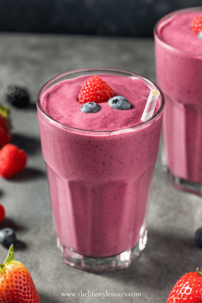 My Go-To Morning Smoothie. Two glasses with bright pink smoothies, raspberries and blueberries on top. Berries scattered on the table.