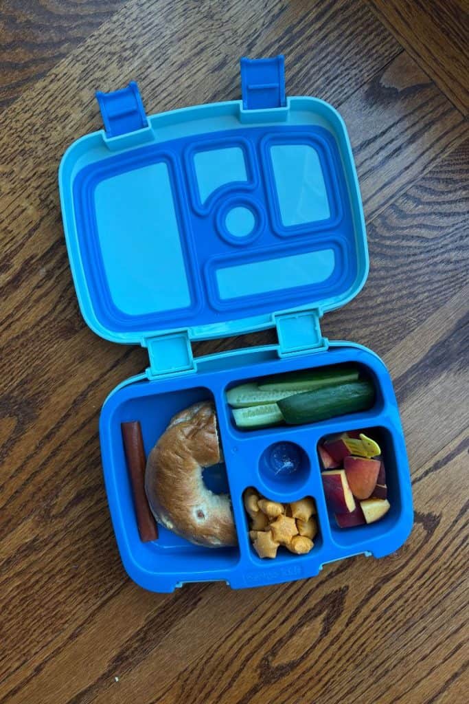 Blue Bentgo box with cucumbers, peaches, food pick, star crackers, Hershey kiss, a half bagel and a small beef stick.
