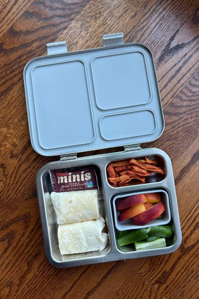Stainless steel Bentgo box with peaches, cucumbers, corn chips, turkey wrap, and mini Larabar.